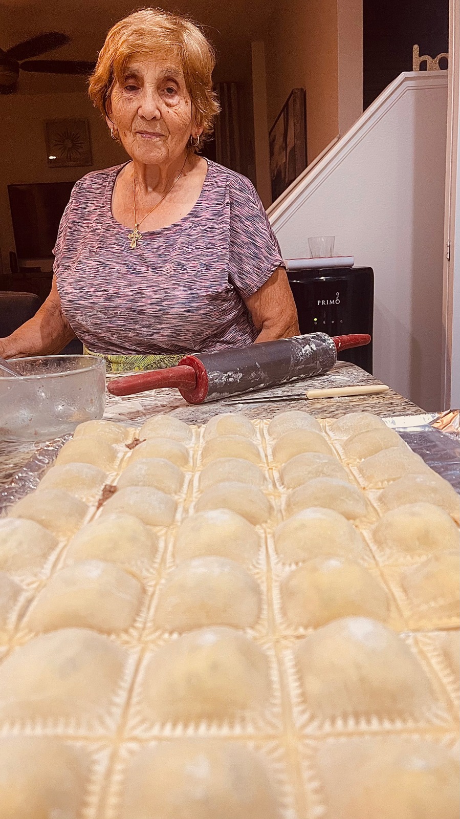 An older woman standing behind a table covered with rows of freshly made ravioli