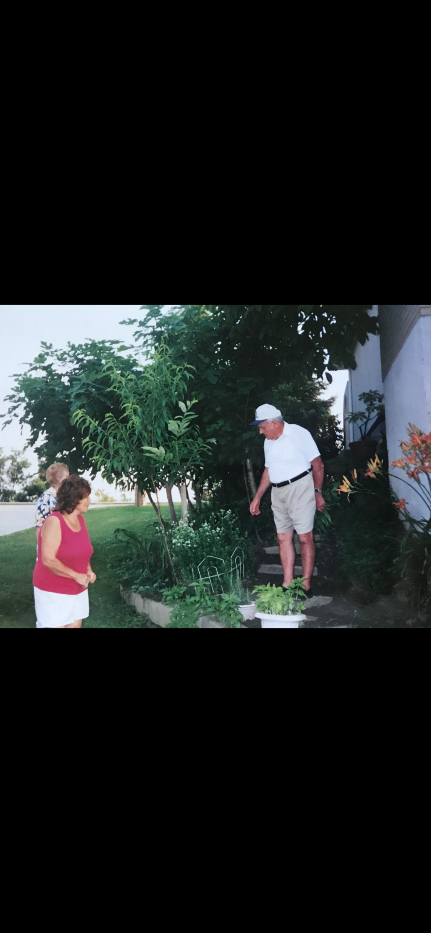 An older man on garden steps examining a plant while a woman in red watches from the lawn