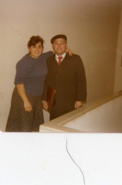 A couple standing arm in arm indoors, the man in a cap and overcoat