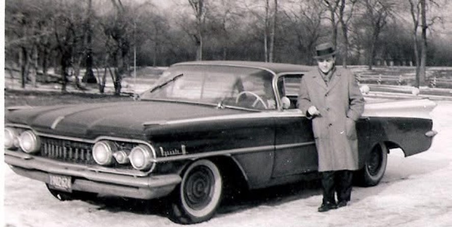 A man in a hat and overcoat standing beside a large car in the snow