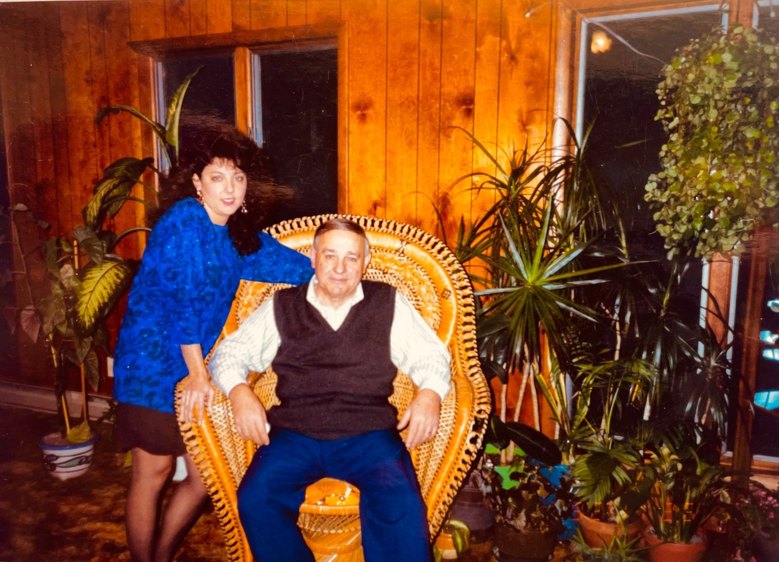 A father seated in a large wicker chair with his daughter beside him, surrounded by plants